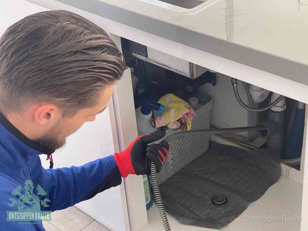 Een monteur voert een flexibele ontstoppingsveer in onder een keukenkastje om een verstopping voorbij de sifon te bereiken in Raalte