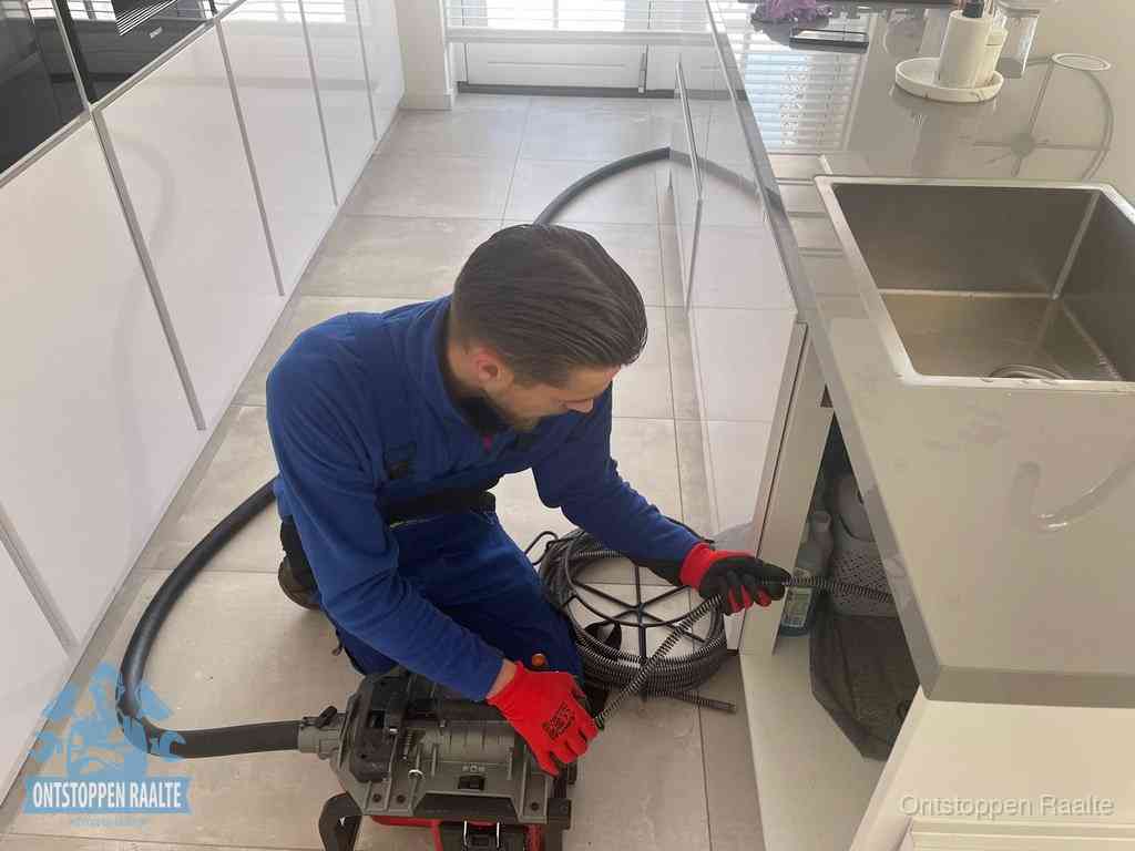 Een monteur ontstopt de keukenafvoer onder de spoelbak met een elektrische veermachine in Raalte