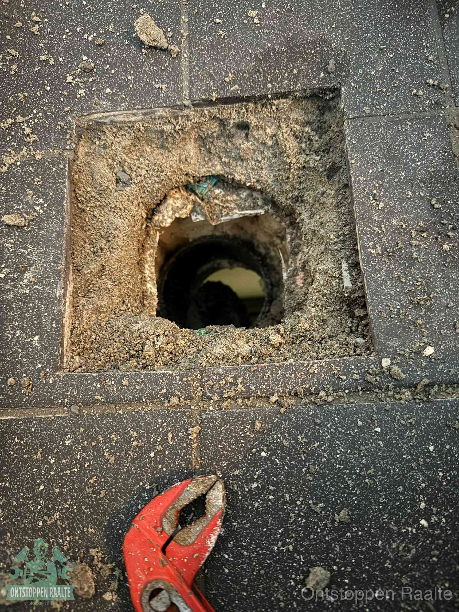Een geopend rioolputje in een betontegelvloer met losgemaakt rooster en een zichtbare ronde afvoerbuis, het achterhalen waar de afvoerverstopping zit in Raalte