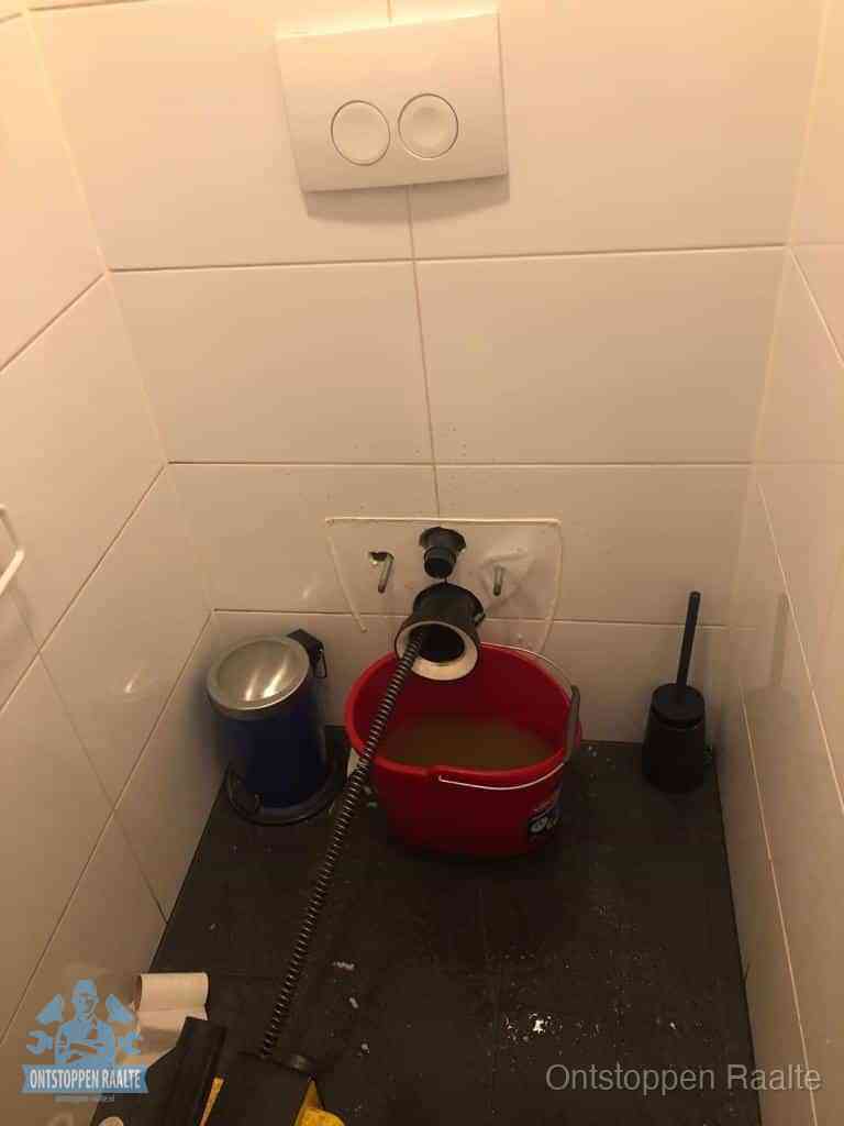 Een ontstopte wand-wc met verwijderde toiletpot waar een ontstoppingsveer in de wanduitgang verdwijnt, het achterhalen waar de verstopping zit in Raalte