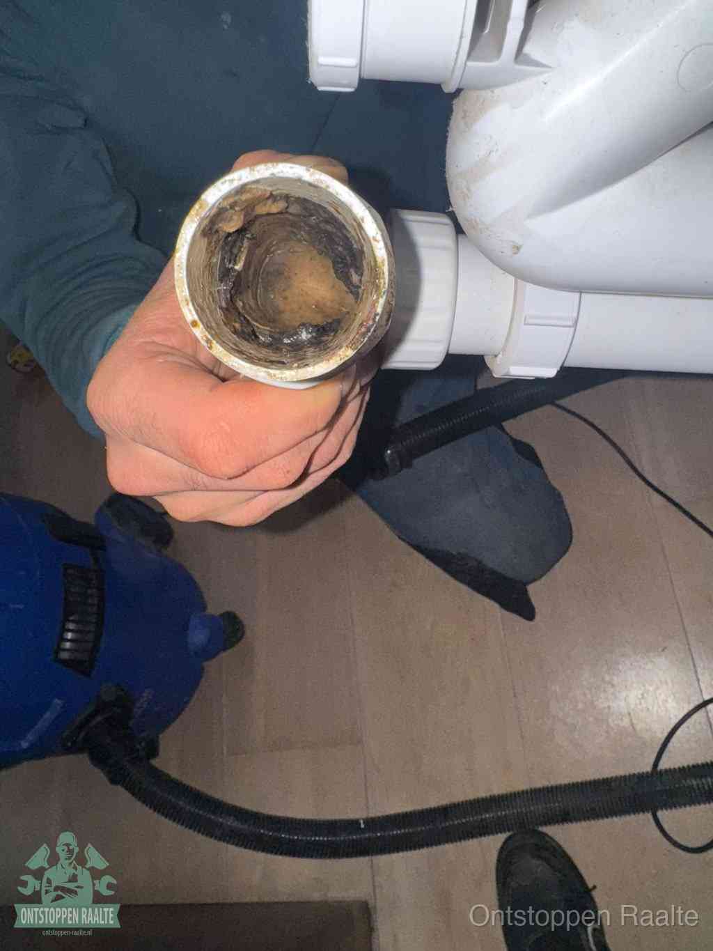 Een hand houdt een losgekoppeld PVC-afvoerbuisstuk vast dat van binnen volledig is dichtgeslibd met vet, vuil en aanslag, de meest voorkomende oorzaak van een verstopte afvoer in Raalte